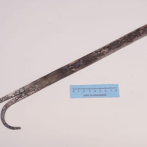 May include: A weathered, dark grey metal tool featuring a long, flat handle that tapers into a sharp, pointed tip and a curved, hook-like extension. A blue paper scale measuring 8 centimetres is placed next to the tool for size reference.