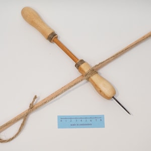 May include: A wooden bow drill tool featuring a light-coloured handle, a central shaft, and a thin metal tip. A piece of twine is wrapped around the shaft and attached to a long wooden rod. A blue ruler shows a scale of 8 centimetres.