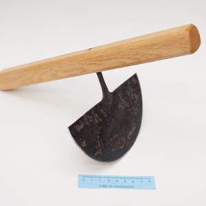 May include: A gardening tool with a wooden handle and a curved, metal blade. The blade is dark grey and has a rough texture. The handle is light brown and has a smooth finish.