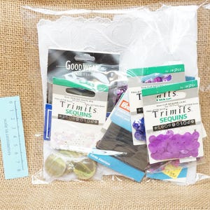 May include: A clear plastic bag containing various craft supplies, including multiple packs of Trimits sequins in purple and white, a white lace fabric piece, and dark thread. A light blue ruler showing 8 centimetres is placed next to the bag.