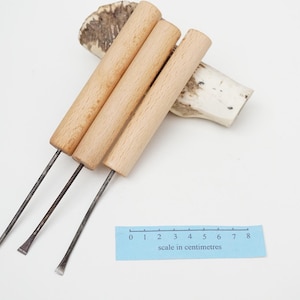 May include: Three metal carving tools with light-coloured wooden cylindrical handles, positioned next to a blue ruler showing a scale in centimetres. The tools feature pointed and flat metal tips, resting against a piece of textured antler.
