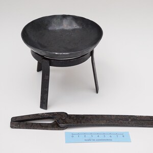 Puede incluir: Un soporte de metal negro con tres patas y un cuenco redondo en la parte superior. Un par de tenazas de metal negro est&aacute;n sobre una superficie blanca junto a una regla marcada en cent&iacute;metros.