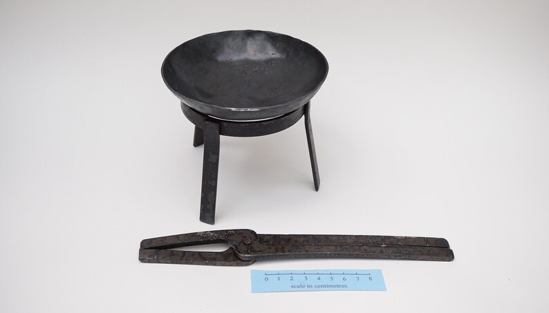 Puede incluir: Un soporte de metal negro con tres patas y un cuenco redondo en la parte superior. Un par de tenazas de metal negro est&aacute;n sobre una superficie blanca junto a una regla marcada en cent&iacute;metros.