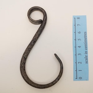 May include: A hand-forged, dark metal hook with a curled top and a curved bottom. The hook is approximately 7 cm long, as indicated by the scale in centimeters next to it. The hook is designed with a rustic, aged appearance.