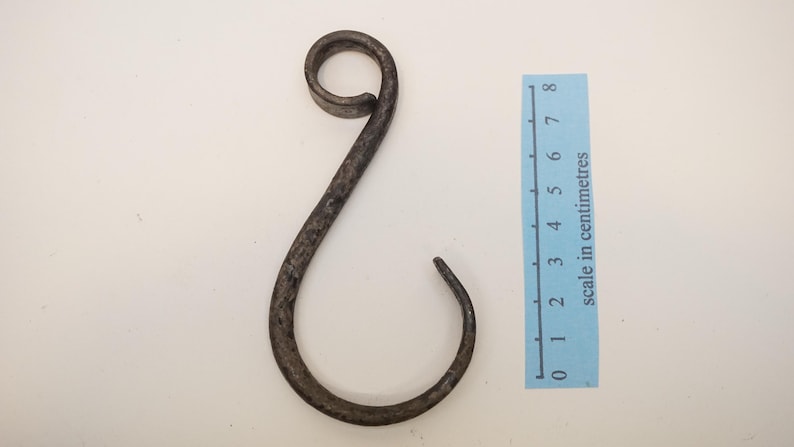 May include: A hand-forged, dark metal hook with a curled top and a curved bottom. The hook is approximately 7 cm long, as indicated by the scale in centimeters next to it. The hook is designed with a rustic, aged appearance.