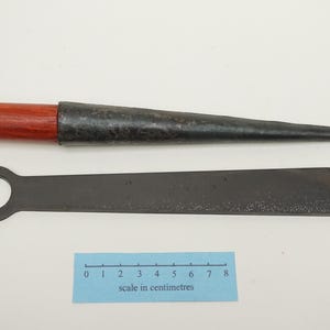 Peut inclure: Un poinçon en métal avec un manche en bois brun rougeâtre et un outil plat en métal séparé avec un trou circulaire à une extrémité. Une échelle bleue mesurant 8 centimètres est placée sous les outils pour indiquer leurs dimensions.