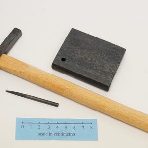 Może przedstawiać: Zestaw narzędzi do obróbki metali, w tym młotek z drewnianym uchwytem, mały kowadło metalowe i stempel metalowy. Do zestawu dołączono linijkę do skalowania.