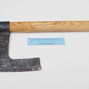 May include: A hand-forged iron axe with a wide, textured blade and a straight, light-coloured wooden handle. A blue paper ruler is positioned below the handle, showing a scale in centimetres from zero to eight, indicating the tool's proportions.