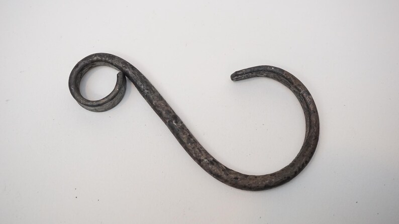 May include: A handcrafted, dark gray metal hook with a distinctive S-shape. The hook features a circular loop on one end and a curved, open hook on the other. The metal has a textured, aged appearance, suggesting a rustic or vintage style.