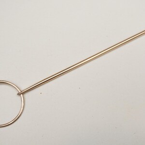 Op de afbeelding: Een goudkleurige hoedenspeld met een cirkelvormige handgreep en een lange, puntige schacht. De handgreep heeft een decoratief knoopontwerp. De speld is bedoeld om hoeden of andere hoofddeksels vast te zetten.