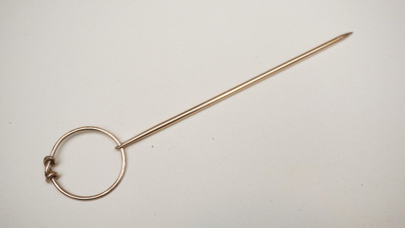 Op de afbeelding: Een goudkleurige hoedenspeld met een cirkelvormige handgreep en een lange, puntige schacht. De handgreep heeft een decoratief knoopontwerp. De speld is bedoeld om hoeden of andere hoofddeksels vast te zetten.