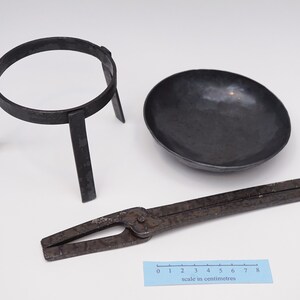 Puede incluir: Un conjunto de tres herramientas de metal vintage para experimentos de qu&iacute;mica. El conjunto incluye un soporte de anillo de metal negro, un taz&oacute;n de metal negro y un par de pinzas de metal negro. Las herramientas est&aacute;n dispuestas sobre una superficie blanca con una regla para la escala.