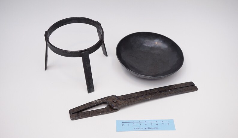 Puede incluir: Un conjunto de tres herramientas de metal vintage para experimentos de qu&iacute;mica. El conjunto incluye un soporte de anillo de metal negro, un taz&oacute;n de metal negro y un par de pinzas de metal negro. Las herramientas est&aacute;n dispuestas sobre una superficie blanca con una regla para la escala.