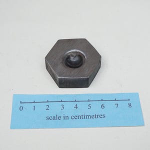 May include: A dark grey, hexagonal metal object with a circular indentation in the centre. A blue ruler with centimetre markings is below the object. The ruler measures up to 8 centimetres.