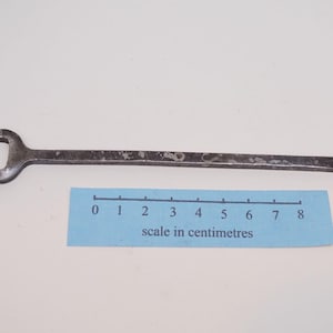 May include: A dark grey, weathered metal two-pronged fork with a long, slender handle and a flattened end. The fork is positioned above a blue paper scale that measures approximately 8 centimetres in length, indicating the tool's size.
