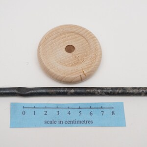 K&ouml;nnte beinhalten: Eine Holzscheibe mit einem Loch in der Mitte und eine schwarze Metallstange, etwa 20 cm lang, mit einer leichten Biegung in der Mitte. Ein Lineal mit Zentimeter-Markierungen ist zur Skalierung enthalten.