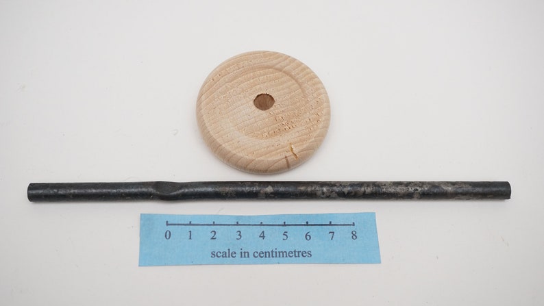 K&ouml;nnte beinhalten: Eine Holzscheibe mit einem Loch in der Mitte und eine schwarze Metallstange, etwa 20 cm lang, mit einer leichten Biegung in der Mitte. Ein Lineal mit Zentimeter-Markierungen ist zur Skalierung enthalten.