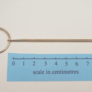 Op de afbeelding: Een goudkleurige hoedenspeld met een cirkelvormige handgreep en een lange, puntige schacht. De speld is ongeveer 7,5 cm lang, zoals aangegeven door de schaal in centimeters op de blauwe liniaal.