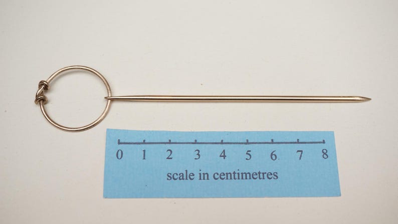Op de afbeelding: Een goudkleurige hoedenspeld met een cirkelvormige handgreep en een lange, puntige schacht. De speld is ongeveer 7,5 cm lang, zoals aangegeven door de schaal in centimeters op de blauwe liniaal.