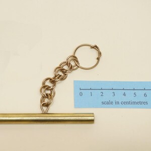 May include: A gold-toned metal toggle bar fastener attached to a short chain of interlocking rings and a circular split ring. A blue paper scale measuring 8 centimetres is placed next to the object to provide a clear visual size reference.