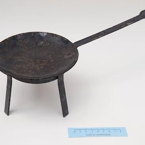 Puede incluir: Una sart&eacute;n de metal negro con un mango largo y tres patas. La sart&eacute;n tiene un di&aacute;metro de unos 18 cm y est&aacute; sobre una superficie blanca.