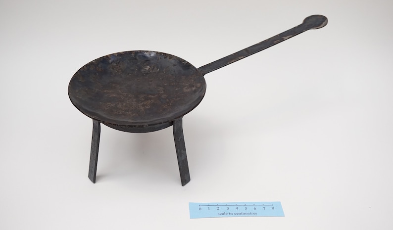 Puede incluir: Una sart&eacute;n de metal negro con un mango largo y tres patas. La sart&eacute;n tiene un di&aacute;metro de unos 18 cm y est&aacute; sobre una superficie blanca.