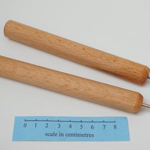 May include: Two cylindrical wooden awls with light brown grain and pointed metal tips. A blue paper ruler is positioned below the tools, showing a length of approximately 8 centimetres for each wooden handle and metal tip assembly.