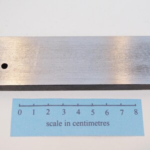 Może przedstawiać: Prostokątny, szczotkowany metalowy przedmiot z małym otworem w pobliżu jednego końca. Niebieska skala w centymetrach znajduje się poniżej. Skala mierzy do 8 cm.