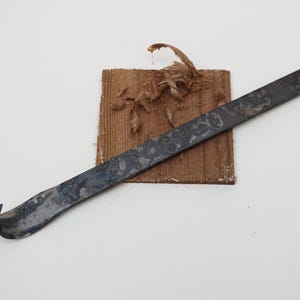 Pode incluir: Uma alavanca de metal escuro com uma extremidade curva repousa sobre um pequeno pedaço de madeira quadrado. A madeira tem uma textura áspera e é coberta com aparas de madeira. O fundo é branco liso.