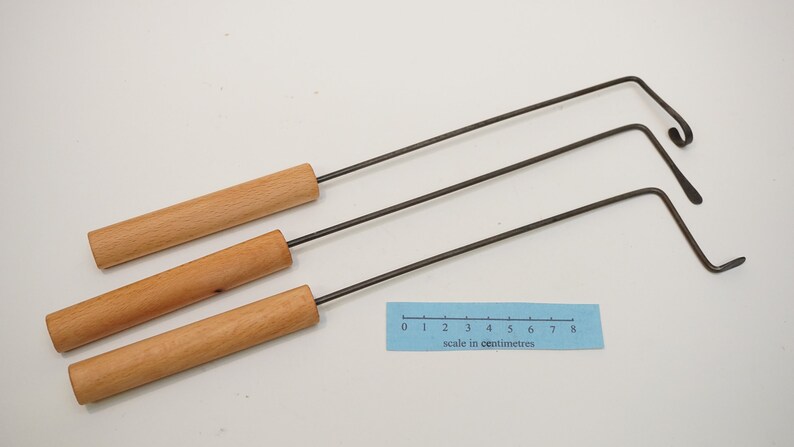 Pu&ograve; includere: Tre strumenti in metallo con manico in legno e un gancio piegato all'estremit&agrave;. Gli strumenti sono disposti su una superficie bianca con un righello che misura 8 centimetri.