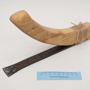 Może przedstawiać: Drewniane narzędzie z zakrzywionym uchwytem i metalowym ostrzem. Uchwyt jest zabezpieczony sznurkiem. W obrazie znajduje się linijka z tekstem "skala w centymetrach".