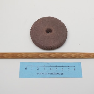 May include: A brown, round, ceramic spinning top with a hole in the center. A wooden stick is lying next to it. A ruler with a scale in centimeters is below the stick.