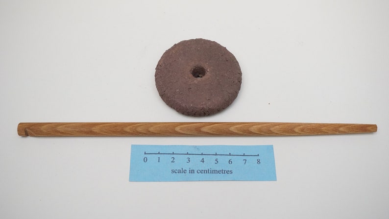May include: A brown, round, ceramic spinning top with a hole in the center. A wooden stick is lying next to it. A ruler with a scale in centimeters is below the stick.