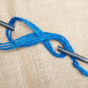 May include: A dark grey metal ring and a pointed metal spike connected by a vibrant blue cord. The cord is looped through the ring and wrapped around the spike, creating a secure knot structure. The metal components have a matte finish.