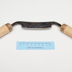 May include: A drawknife featuring a dark, tempered metal blade with a mottled finish and two light-coloured wooden handles. A blue paper ruler is positioned below the blade, showing a measurement scale in centimetres from zero to eight centimetres.