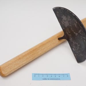 May include: A gardening tool with a curved, metal blade and a wooden handle. The blade is dark grey and has a rough texture. The handle is light brown and measures approximately 20 cm in length. The tool is on a white surface with a ruler that reads "scale in centimetres".