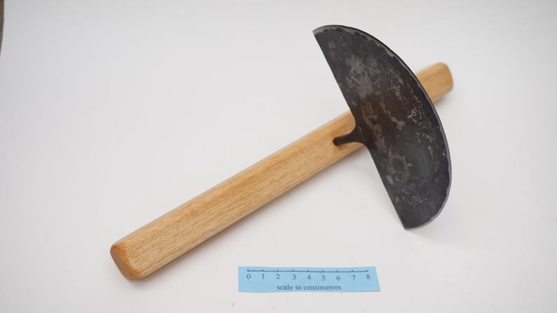 May include: A gardening tool with a curved, metal blade and a wooden handle. The blade is dark grey and has a rough texture. The handle is light brown and measures approximately 20 cm in length. The tool is on a white surface with a ruler that reads "scale in centimetres".