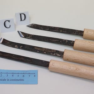 May include: Four wood carving chisels with light-coloured wooden handles and dark metal blades of varying shapes. Each tool is labelled with a white card marked A, B, C, or D. A blue ruler shows a scale in centimetres from 0 to 8 centimetres.