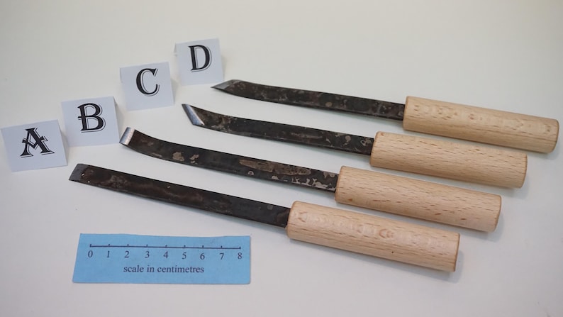 Puede incluir: Cuatro formones para madera con mangos de madera clara y hojas de metal oscuro de diversas formas. Cada herramienta est&aacute; etiquetada con una tarjeta blanca marcada con A, B, C o D. Una regla azul muestra una escala de 0 a 8 cent&iacute;metros.
