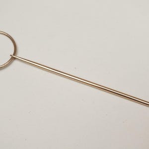 Op de afbeelding: Een goudkleurige hoedenspeld met een cirkelvormige bovenkant en een lange, puntige speld. De cirkelvormige bovenkant heeft een decoratief knoopontwerp. De speld is ongeveer 13 cm lang.