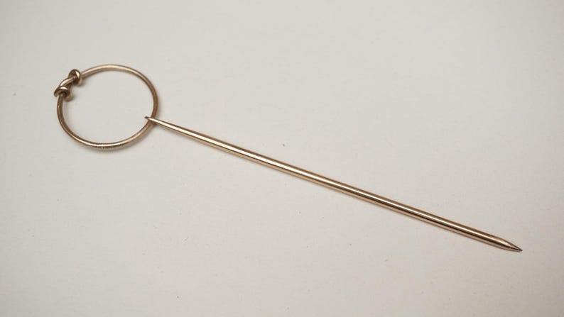 Op de afbeelding: Een goudkleurige hoedenspeld met een cirkelvormige bovenkant en een lange, puntige speld. De cirkelvormige bovenkant heeft een decoratief knoopontwerp. De speld is ongeveer 13 cm lang.