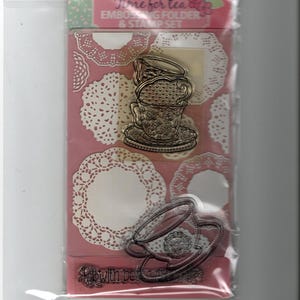 May include: A clear stamp set with a stacked teacup design, the words "Time for tea" and "Embossing folder & stamp set". The background is pink with white doily patterns. The set is in a clear plastic package.