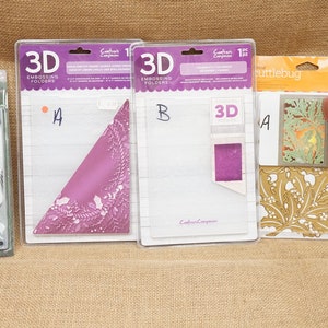 May include: Five different embossing folders for crafting. The folders are packaged in clear plastic with white and purple lettering. The folders feature a variety of designs, including a snowflake, a floral pattern, and a geometric design. The folders are labelled with the letters A and B.