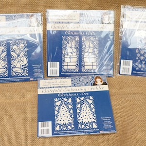 May include: A collection of Gatefold Embossing Folders in plastic wrapping. The folders feature white designs on a blue background, including bells, Christmas gifts, snowflakes, and Christmas trees. They are on a brown burlap surface.