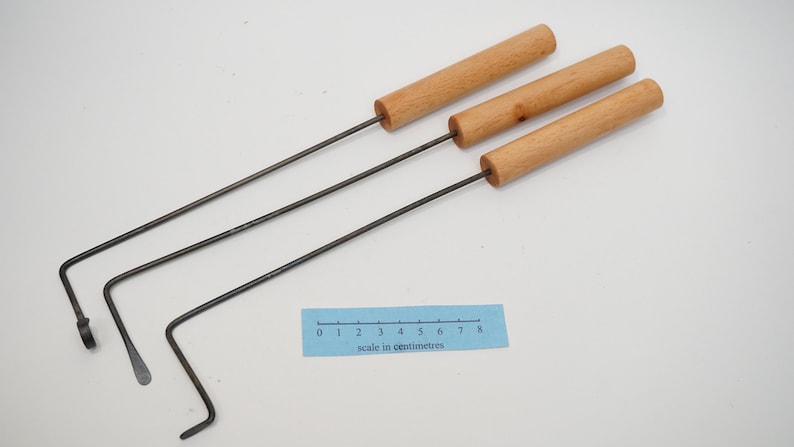 Pu&ograve; includere: Tre strumenti in metallo con manici in legno e un gancio piegato all'estremit&agrave;. Gli strumenti sono disposti su una superficie bianca con un righello che misura 8 centimetri.