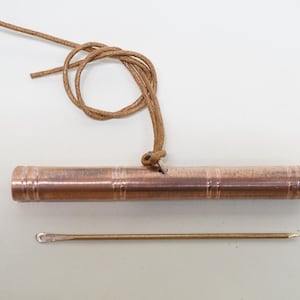 Könnte beinhalten: Ein Kupferrohr mit einem braunen Lederband daran. Eine dünne Metallnadel liegt neben dem Rohr.