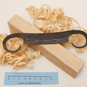 Può includere: Un coltello da trazione forgiato a mano con una lama scura e strutturata e manici ricurvi, appoggiato su un blocco di legno. Il coltello è circondato da trucioli di legno, con un righello blu che mostra i centimetri.