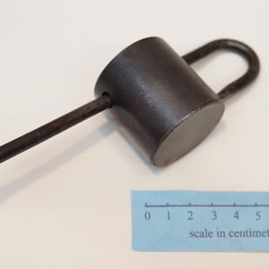 Peut inclure: Un cadenas cylindrique en métal brun foncé avec une anse en forme de U et une longue clé fine à bout plat insérée sur le côté. Une échelle en papier bleu avec des graduations de 0 à 8 centimètres est placée sous l'objet pour référence.