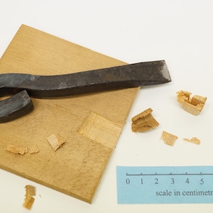 Op de afbeelding: Een gebogen metalen gereedschap met een houten handvat, liggend op een houten oppervlak. Er zijn houtkrullen rondom het gereedschap verspreid. Een liniaal met een schaalverdeling in centimeters is zichtbaar aan de onderkant van de afbeelding.