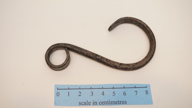 May include: A rustic, dark metal hook with a curved design. The hook is approximately 8 inches long, with a circular loop on one end and a curved hook on the other. The hook is likely for hanging items.